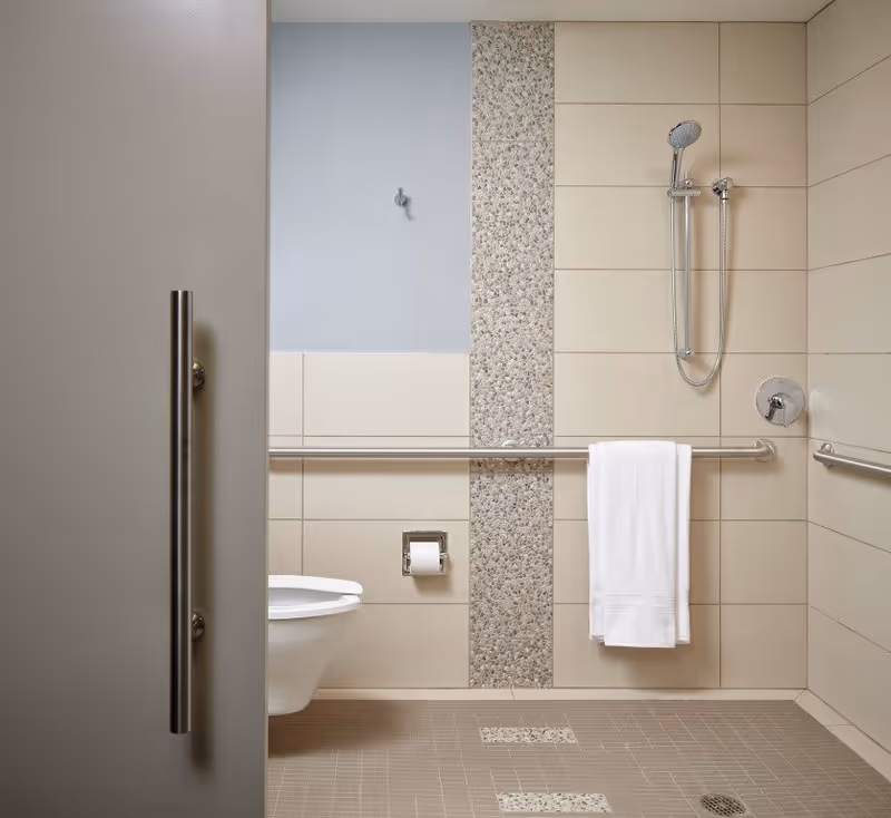 A modern, accessible bathroom with beige tiled walls and floor, featuring a wall-mounted toilet, a stainless steel grab bar with a white towel hanging on it, a handheld showerhead, and a vertical strip of pebble-textured tiles on the wall.