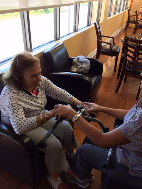 An elderly woman sitting in a black armchair holding hands with a person sitting across from her in a room with wooden floors and large windows. The person across is wearing scrubs and holding a walker. There are more chairs and tables visible in the background.