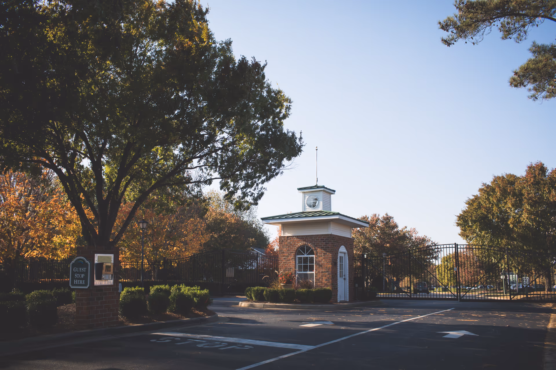 Entrance gate to a facility with a small brick guardhouse featuring a clock on top, surrounded by trees with autumn foliage and a sign that says 'Guest Stop Here'.