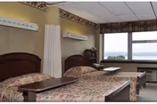 A shared senior care room with two beds, a privacy curtain between them, bedside tables, and a large window overlooking water.