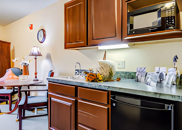 Small kitchen area with dark wooden cabinets, a countertop and sink, microwave above, and a dining table with chairs in the background.