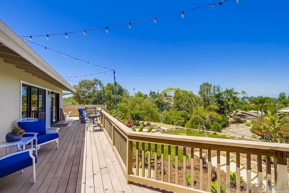A spacious wooden deck attached to a building with blue cushioned chairs and a small table. String lights are hung above the deck. The deck overlooks a landscaped garden with various trees and plants under a clear blue sky.