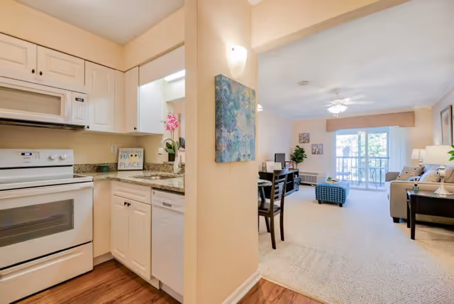 View of a small kitchen area with white cabinets, a white stove, microwave, and dishwasher. The kitchen has granite countertops and a decorative sign that reads 'This is my happy place.' Adjacent to the kitchen is a living room with beige carpet, a ceiling fan, a sofa, a blue ottoman, a TV on a stand, and sliding glass doors leading to a balcony.