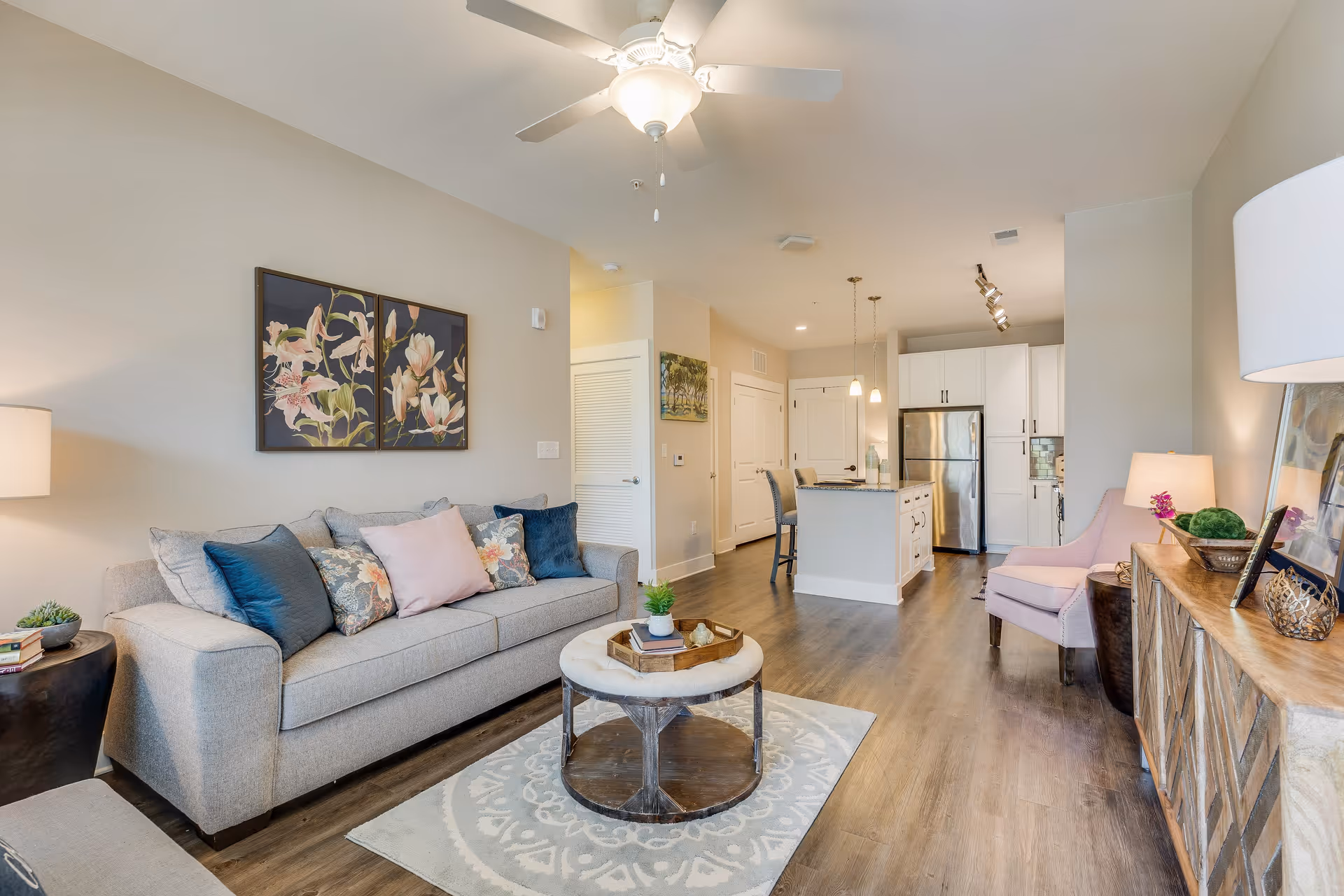 A bright and modern living room with a gray sofa adorned with multiple decorative pillows, a round coffee table on a patterned rug, and a wooden sideboard with a lamp and decorative items. The room opens into a kitchen area with white cabinets, stainless steel refrigerator, and an island with bar stools. The walls are light-colored, and the floor is wooden. A ceiling fan with light is visible above.