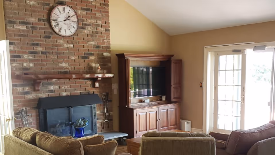 A cozy living room with a brick fireplace on the left wall, a large clock above the mantel, and a wooden entertainment center with a TV. There are several cushioned chairs and sofas arranged around a wooden coffee table. Sunlight streams in through glass double doors on the right side.