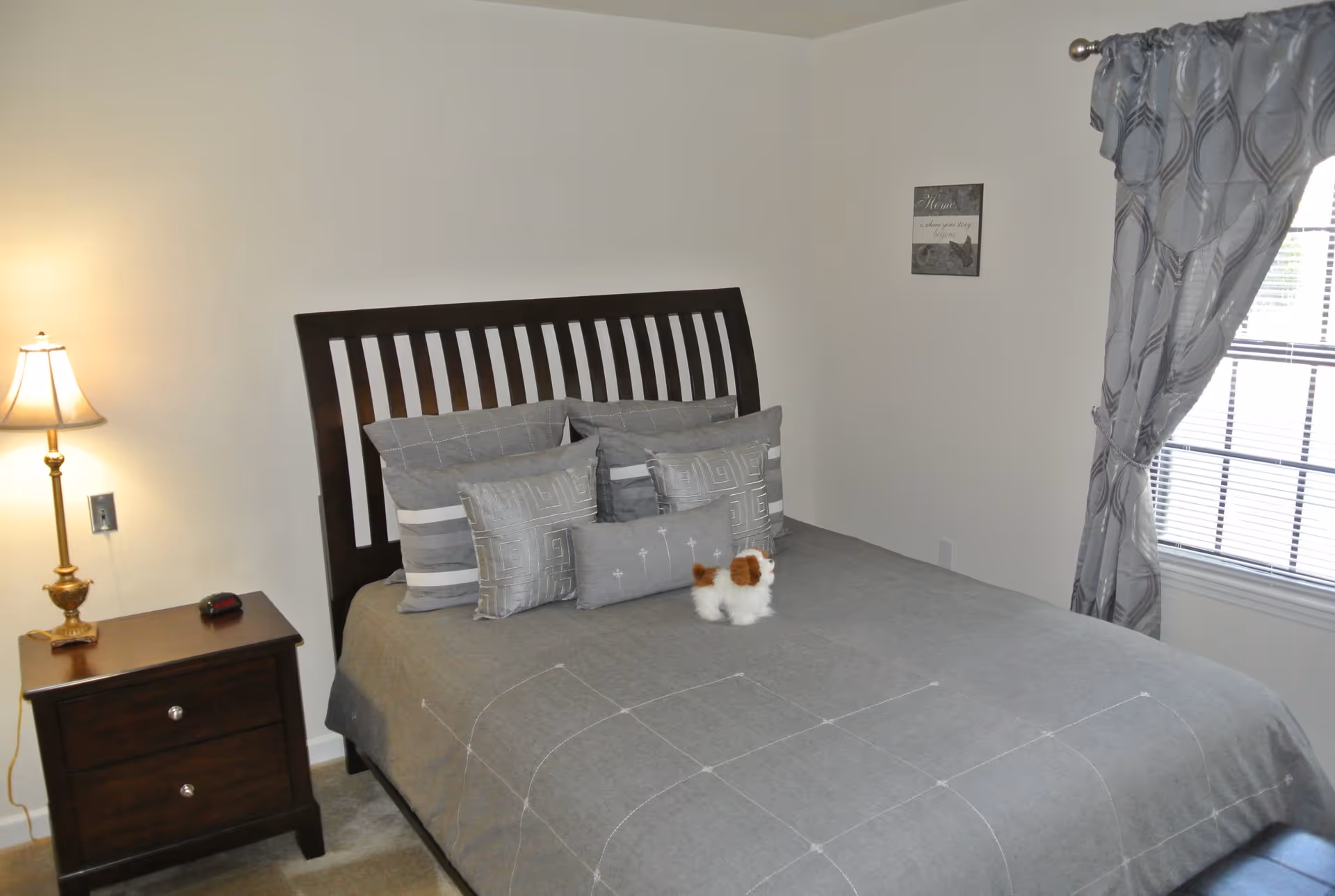 A bedroom with a dark wooden bed frame and a neatly made bed with gray bedding and multiple pillows. A small stuffed dog toy is placed on the bed. To the left of the bed is a wooden nightstand with a lamp and a digital alarm clock. A window with gray patterned curtains is on the right wall, and a small framed sign hangs on the wall above the bed.