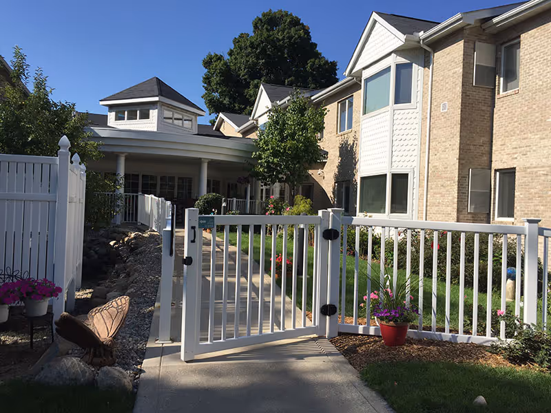 Gated white fence and walkway leading to the front entrance of a brick senior living building with landscaping and potted flowers.