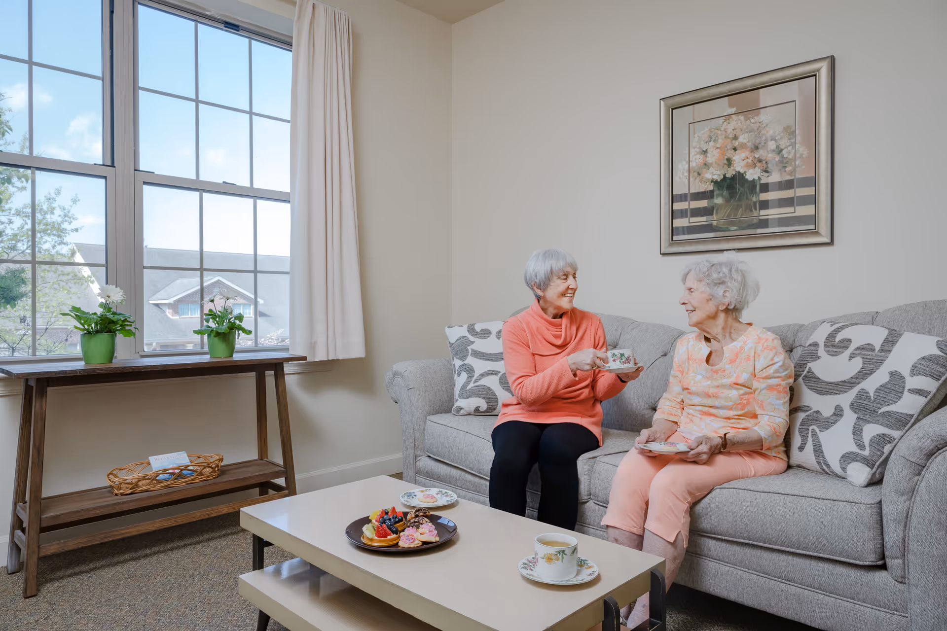 Two elderly women sitting on a gray couch in a bright living room, enjoying tea and pastries. The room has a large window with white curtains, a wooden table with two potted plants, and a framed floral painting on the wall.