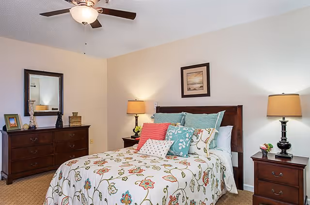 A cozy bedroom with a wooden bed frame and a bed dressed in a floral patterned comforter and multiple colorful pillows. There are two wooden nightstands on either side of the bed, each with a lamp. A wooden dresser with a mirror and decorative items is positioned against the wall. The walls are light-colored, and a framed picture hangs above the bed.
