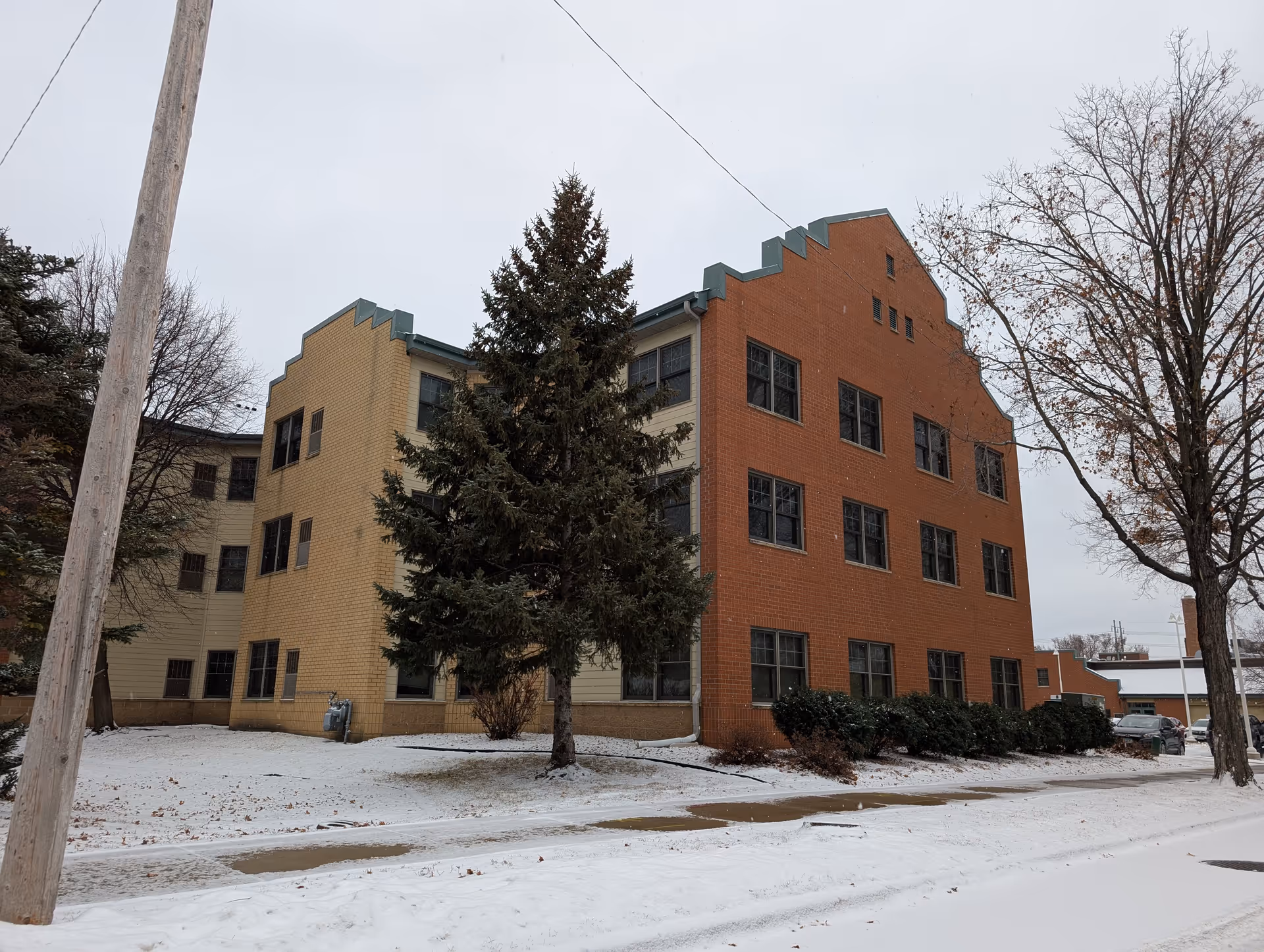 Exterior view of a three-story brick building with a stepped roofline, surrounded by snow-covered ground, leafless trees, and a sidewalk. The building has multiple windows and is partially obscured by a large evergreen tree in the foreground.