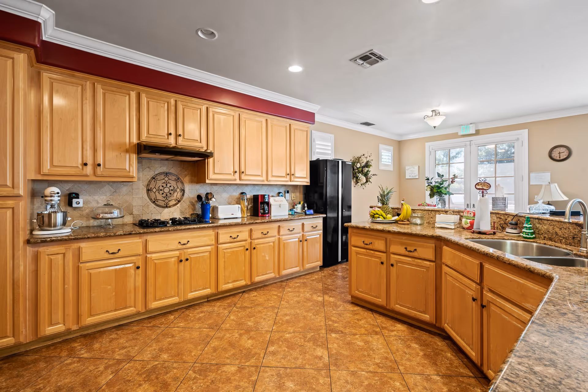 A spacious kitchen with wooden cabinets, granite countertops, and a tiled floor. The kitchen features a black refrigerator, a gas stove with a decorative tile backsplash, a double sink, and various kitchen appliances and utensils on the counters. There are windows and a glass door letting in natural light, with some decorative plants and a clock on the wall.