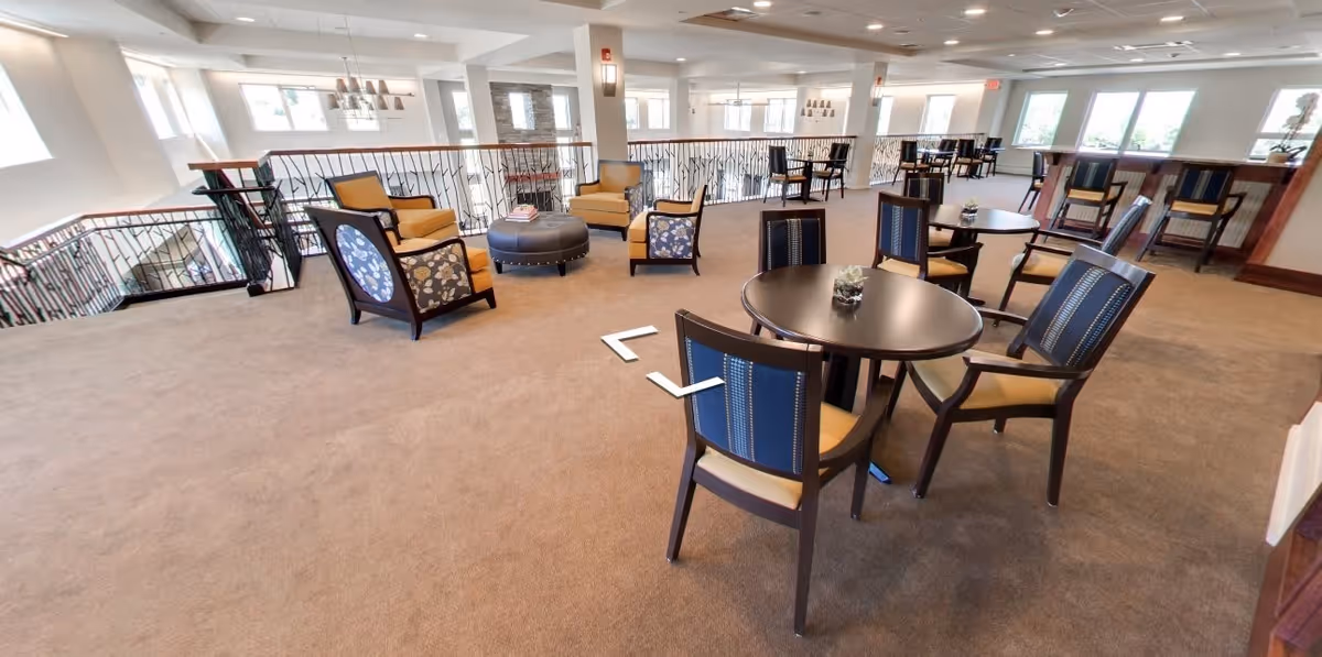 Spacious indoor common area with multiple seating arrangements including round tables with chairs and cushioned armchairs around a central ottoman. The area has carpeted floors, large windows allowing natural light, and a railing overlooking a lower level.