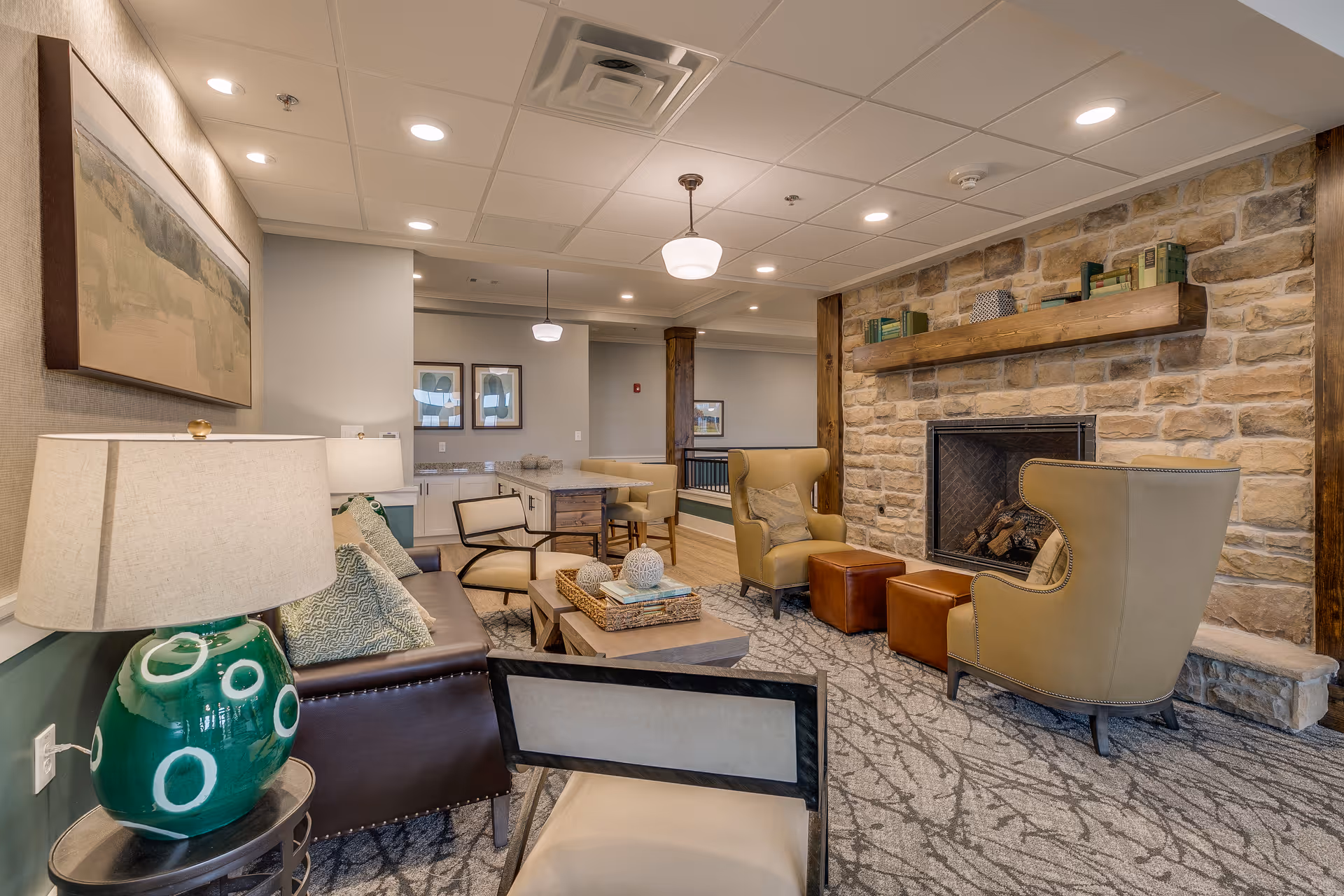 A cozy living room area in a senior living facility featuring a stone fireplace with a wooden mantel decorated with books and ornaments. The room has two beige wingback chairs with brown ottomans, a brown leather sofa with patterned cushions, a coffee table with decorative items, and a green lamp on a side table. The walls are painted in neutral tones with framed artwork, and the ceiling has recessed lighting and pendant lights.
