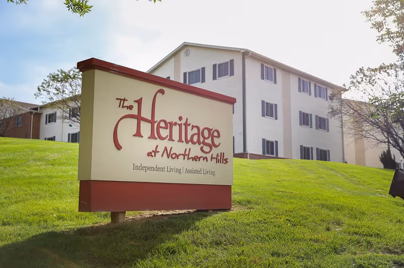 A large sign on a grassy hill reads 'The Heritage at Northern Hills Independent Living | Assisted Living' with a multi-story residential building in the background under a partly cloudy sky.