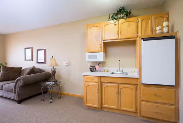 A small kitchenette area with wooden cabinets, a white microwave, a sink, and a refrigerator. To the left, there is a brown sofa with cushions, a glass side table with a lamp, and two framed pictures on the beige wall.