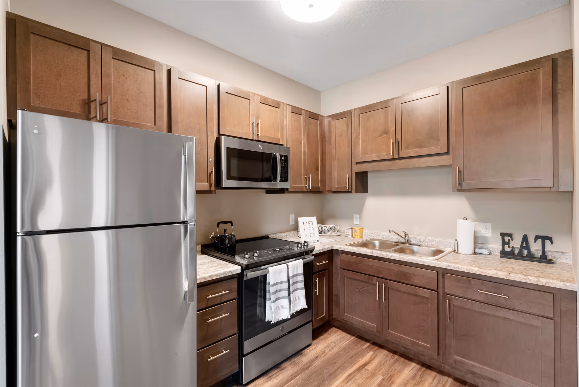 Modern kitchen with stainless steel refrigerator, stove, and microwave. The kitchen features wooden cabinets, a double sink, and a countertop with a paper towel holder and decorative letters spelling 'EAT'.