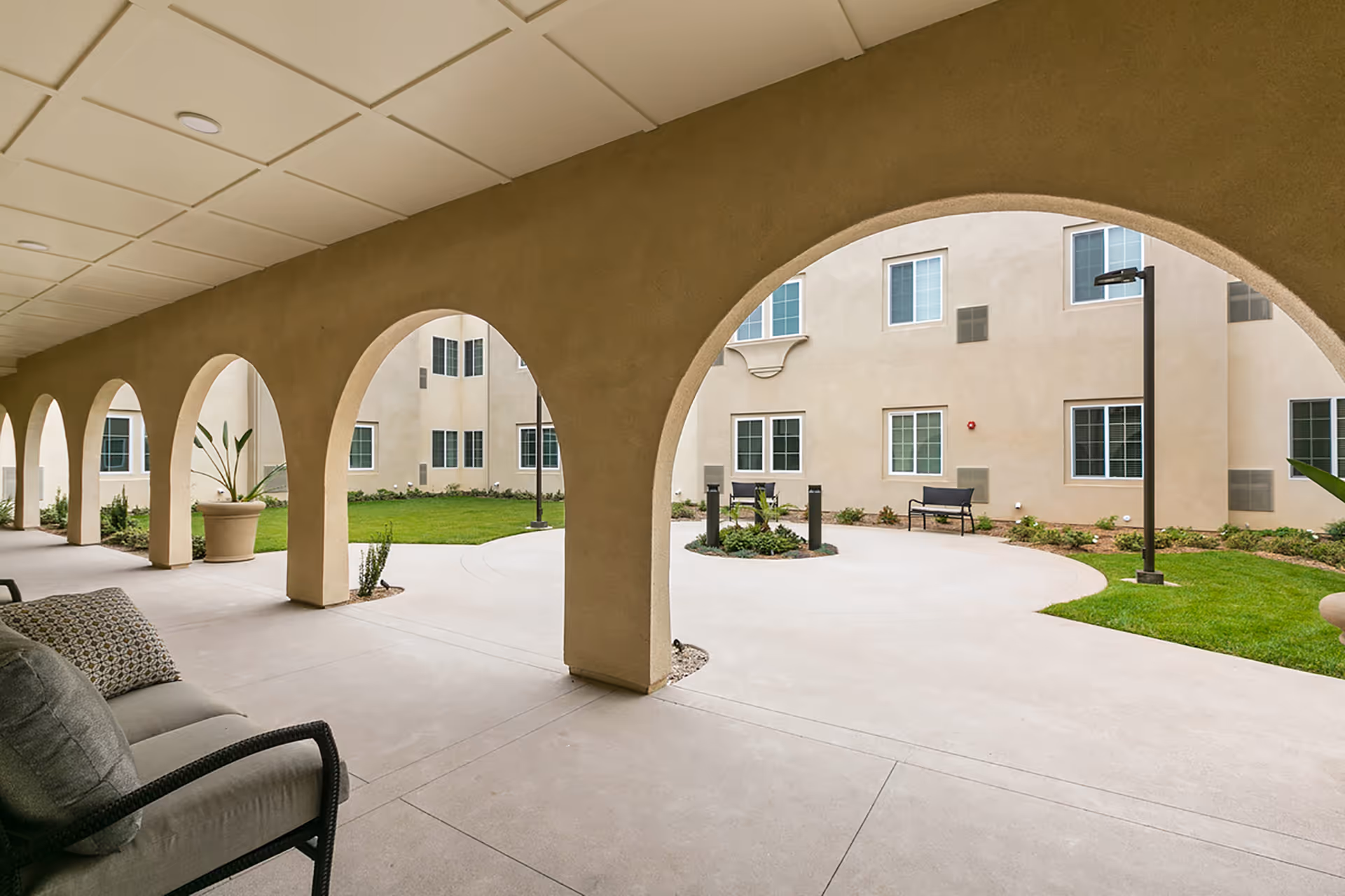 Covered arched walkway opening onto a landscaped courtyard with benches and surrounding building windows.