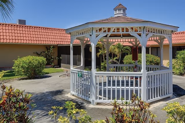 A white wooden gazebo with a shingled roof situated in a courtyard surrounded by greenery and a building with a red tiled roof under a clear blue sky.