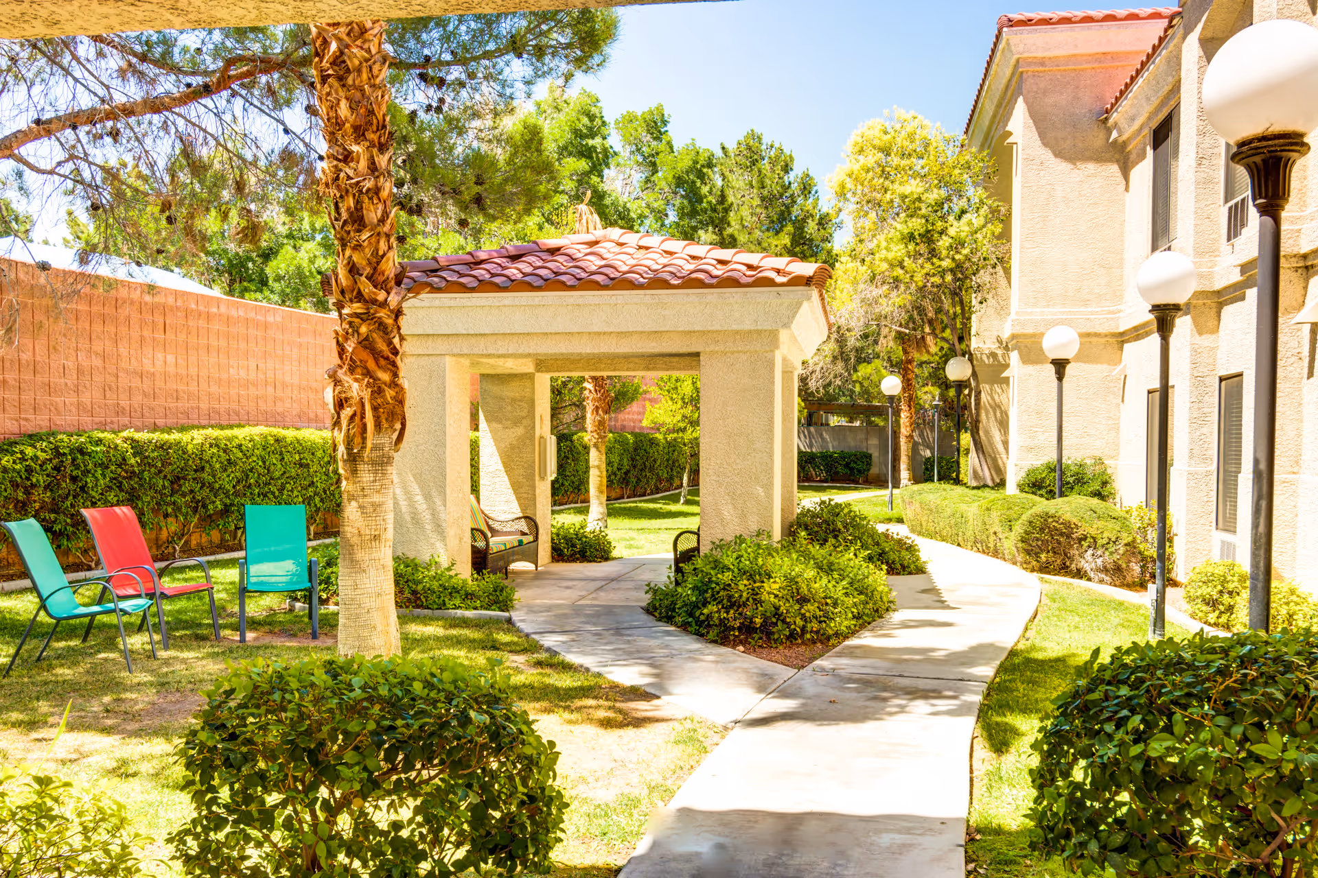 Outdoor garden area at Escalante at the Lakes featuring a paved walkway, green bushes, palm trees, a small covered seating area with benches, and colorful chairs on the grass. The scene is bright and sunny with a building on the right side and lamp posts lining the path.