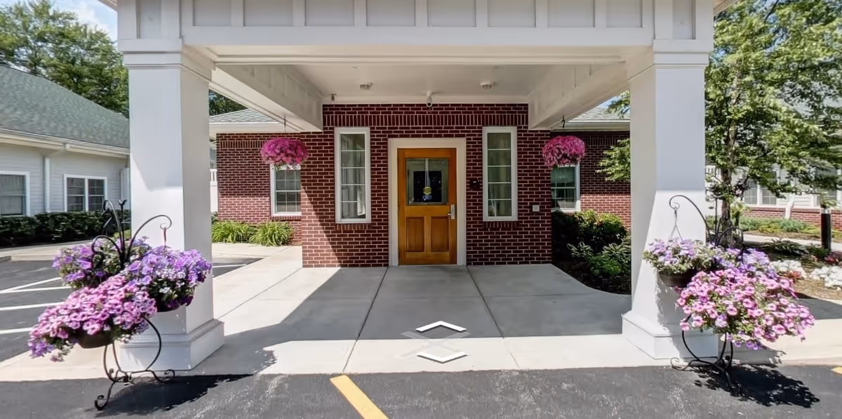 Entrance of a senior living facility with a covered porch supported by white columns. There are hanging baskets and planters with purple flowers on either side of the entrance. The building has red brick walls and a wooden door with glass panels.
