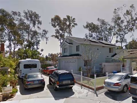Residential-style building with multiple parked cars in a driveway and a fenced front yard