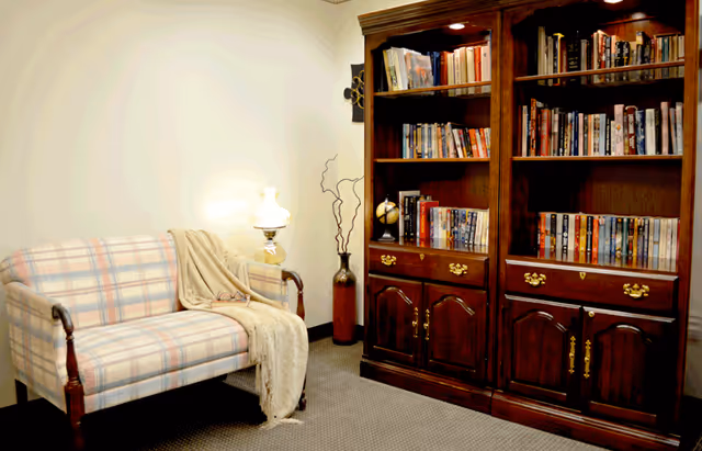 A cozy interior room featuring a plaid upholstered loveseat with wooden armrests, draped with a beige throw blanket and a pair of reading glasses resting on it. Next to the loveseat is a small table lamp with a white shade, and behind it, a tall decorative vase with dried branches. On the right side, there is a large wooden bookshelf filled with books and a small globe, with cabinets and drawers at the bottom.