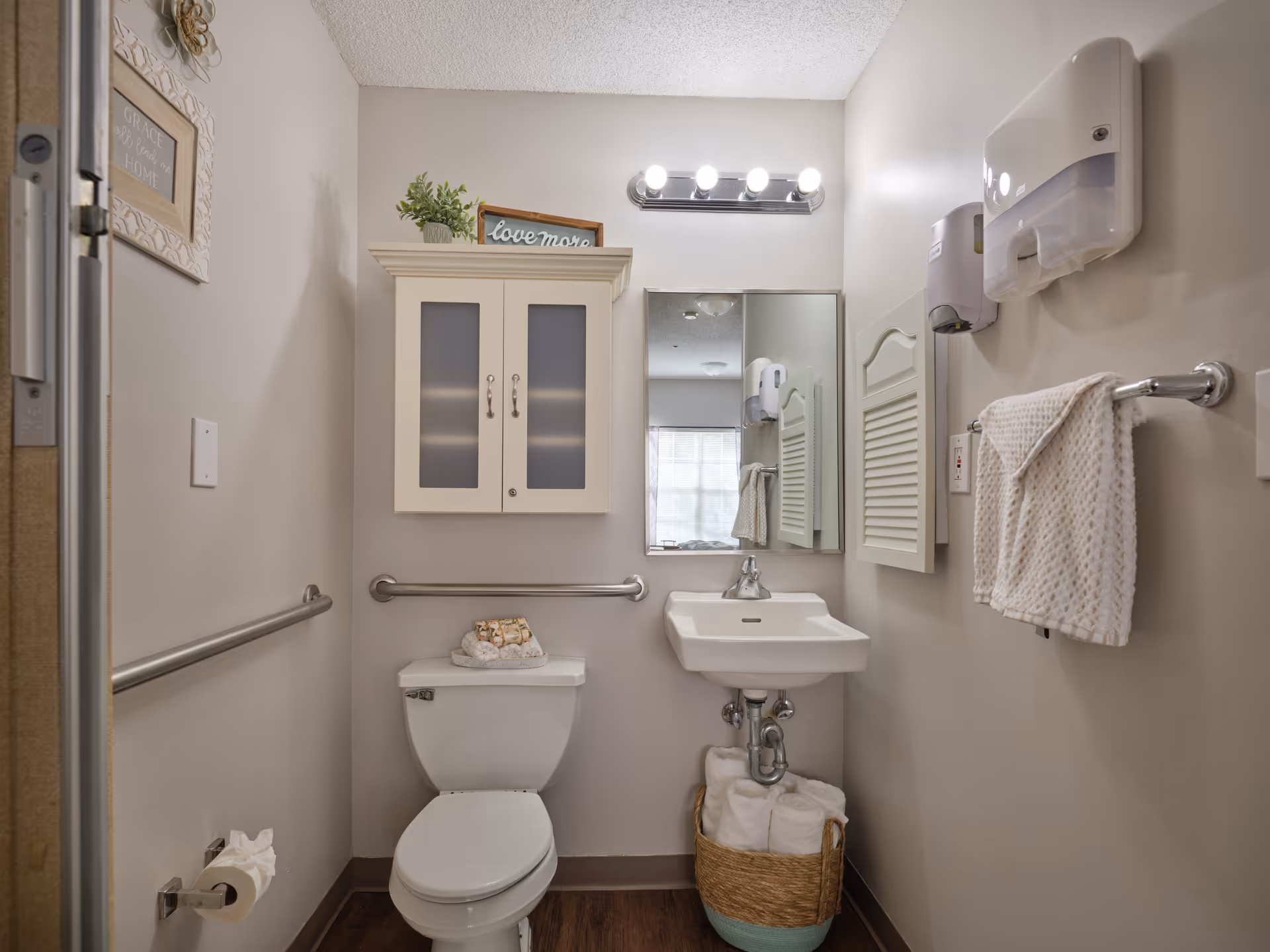 A small, clean bathroom with a white toilet and a white sink. Above the toilet is a cabinet with frosted glass doors and a small plant and decorative sign on top. A mirror with lights above it is mounted on the wall above the sink. There are grab bars on the walls near the toilet and a towel rack with a white towel. A basket with rolled white towels is placed under the sink. The walls are painted light beige and the floor is wood.