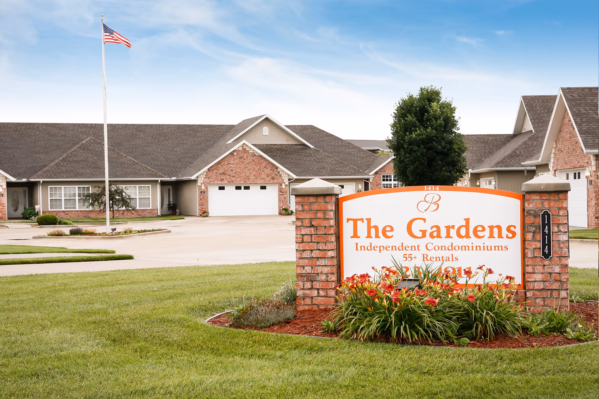 Entrance sign for The Gardens Independent Condominiums 55+ Rentals with brick pillars and flowers in front, residential buildings with garages and an American flag on a flagpole in the background under a blue sky.