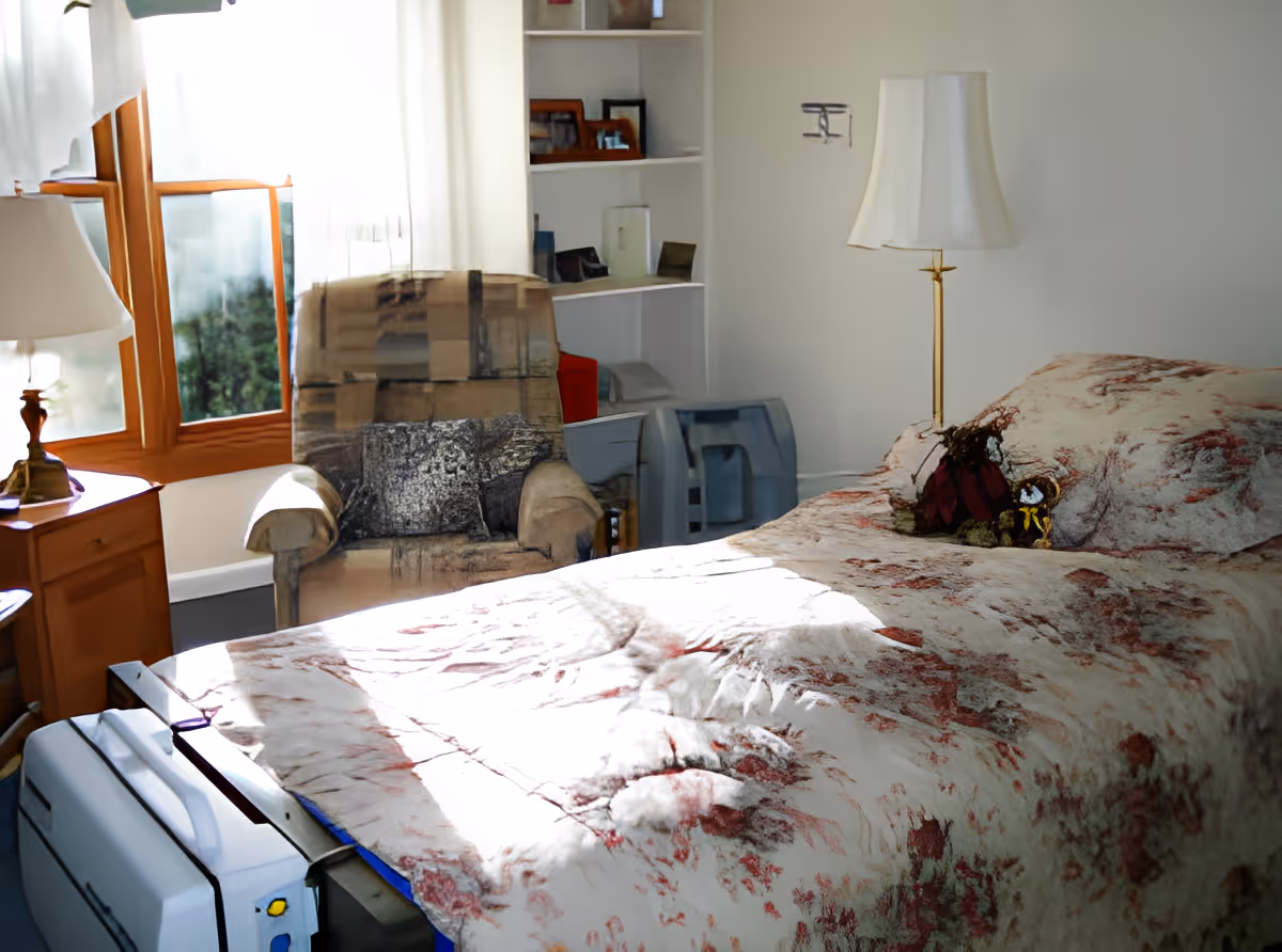 A cozy bedroom with a single bed covered in a floral-patterned quilt. Next to the bed is a wooden nightstand with a lamp. There is a cushioned armchair near a window with sheer white curtains, allowing natural light to fill the room. A white shelving unit with various items is visible in the background.