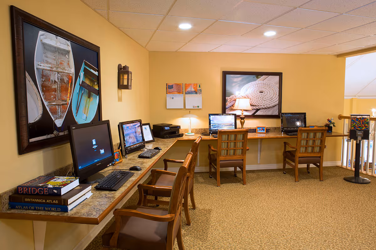 A computer workstation area in a senior living facility with four desktop computers on a long counter against a yellow wall. There are wooden chairs with cushions in front of each computer. The wall is decorated with framed pictures of boats and a coiled rope, a calendar, and a small lamp. Books are stacked on the counter, and the floor is carpeted.