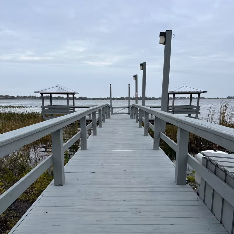 Wooden boardwalk pier with railings leading out to a lake, flanked by small covered seating areas under an overcast sky.