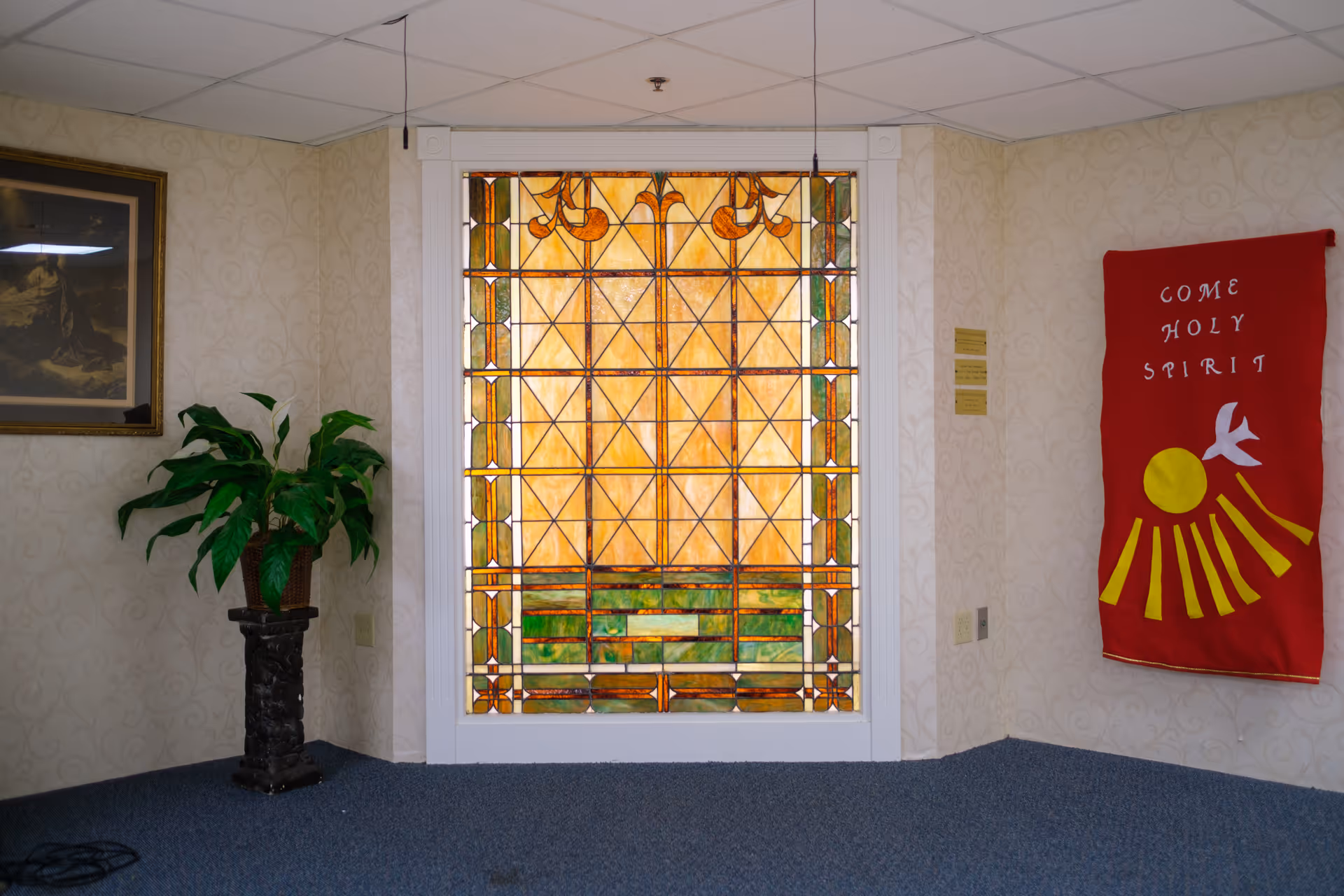 Interior corner of a room featuring a large stained glass window with geometric patterns in yellow, green, and orange hues. To the left of the window is a potted plant on a dark pedestal and a framed picture on the wall. To the right of the window hangs a red banner with the words 'COME HOLY SPIRIT' and a white dove above a yellow sun with rays. The room has light patterned wallpaper and blue carpeted floor.