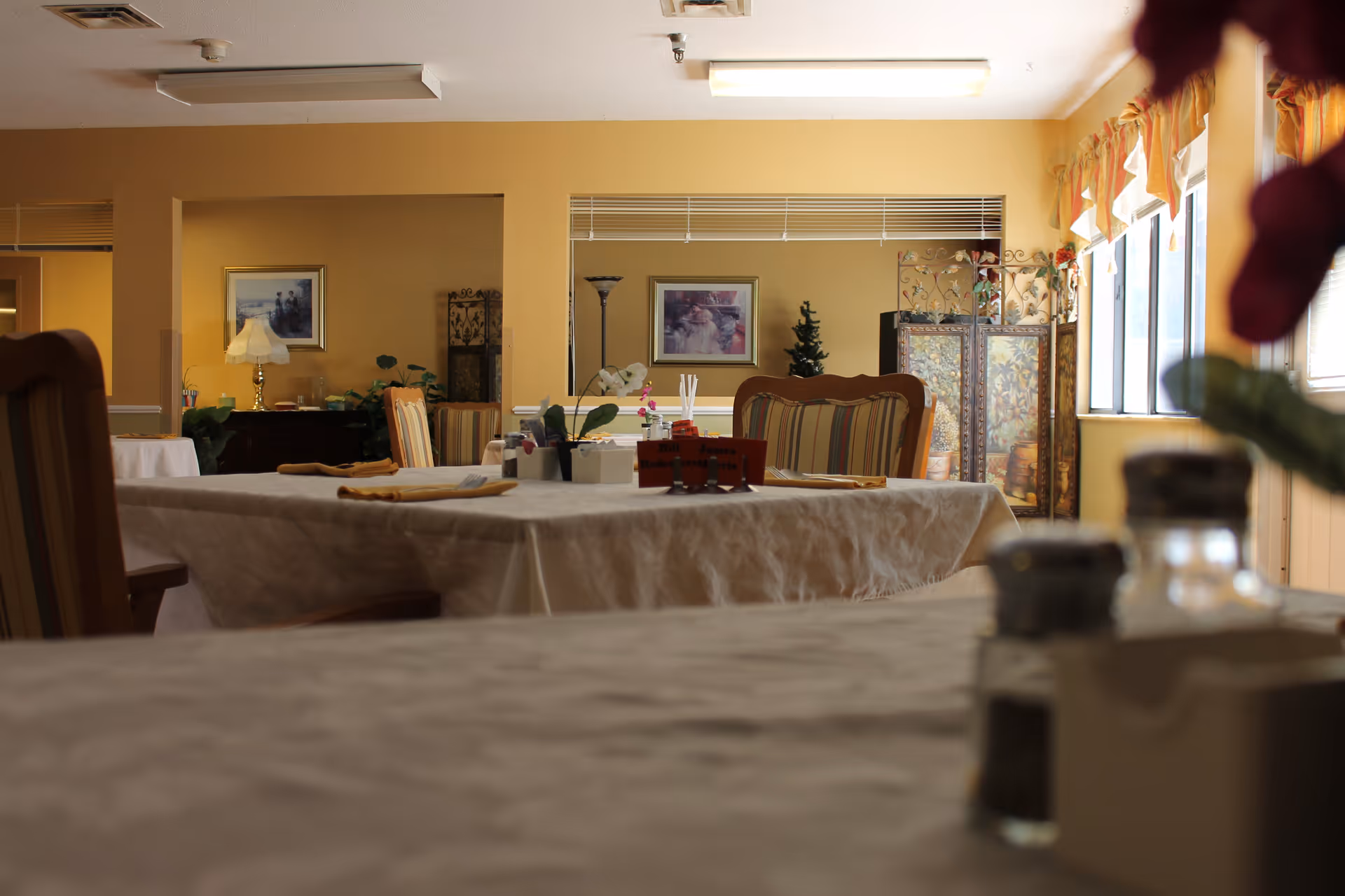 Communal dining room with tablecloth-covered tables, chairs, place settings, and windows along the right side.