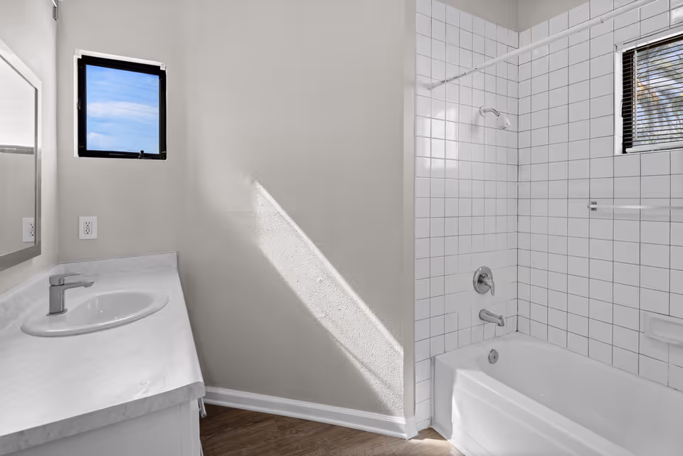 A clean bathroom with a white sink and countertop on the left, a mirror above the sink, and a bathtub with white tiled walls and a showerhead on the right. There are two small windows with black frames allowing natural light into the room.