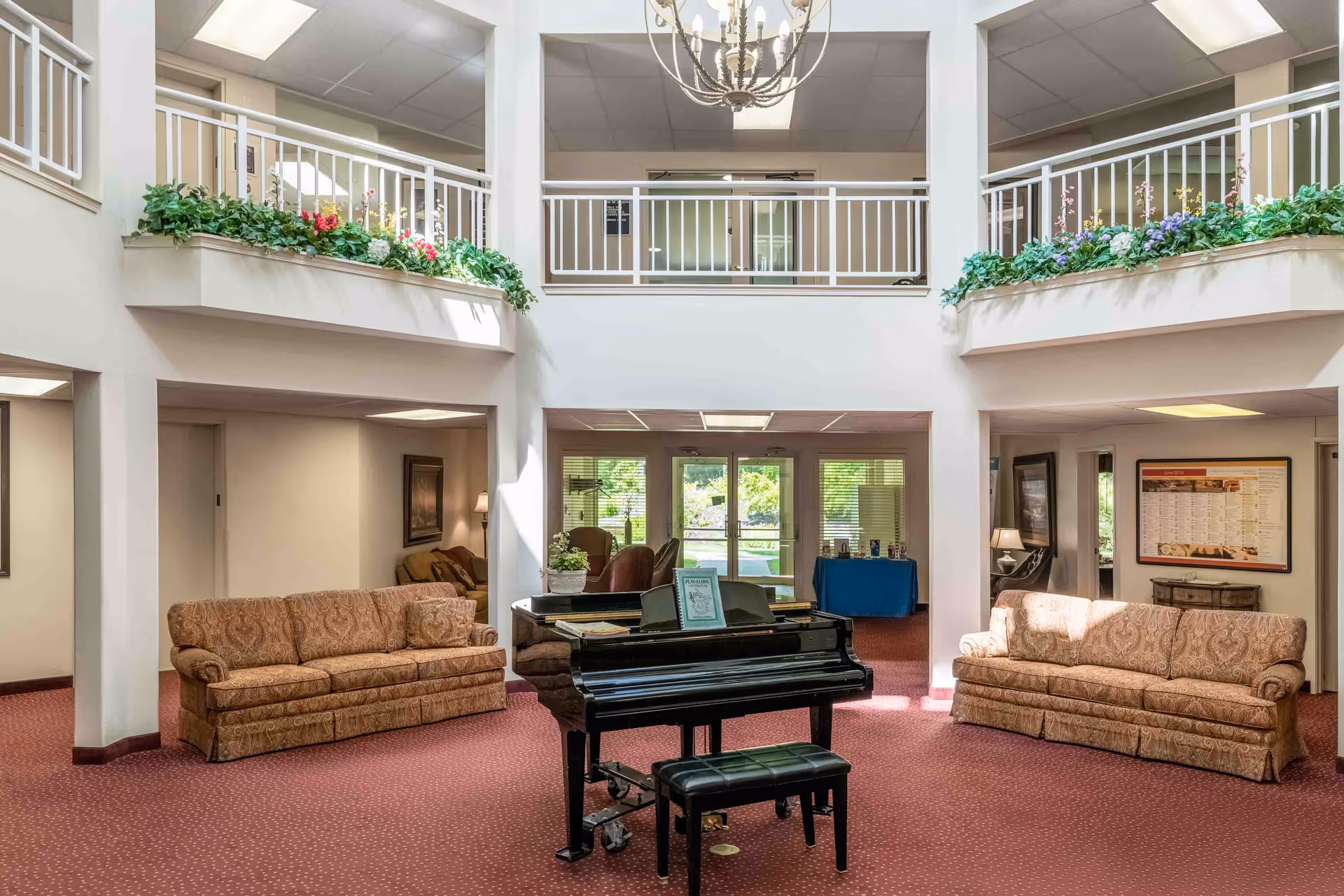 A spacious senior living facility common area with two patterned sofas facing each other on a red carpet. A black grand piano with a matching bench is centered in the room. The area has high ceilings with a chandelier and a second-floor balcony adorned with green plants and flowers. Large windows and glass doors at the back let in natural light and provide a view of the outdoor greenery.
