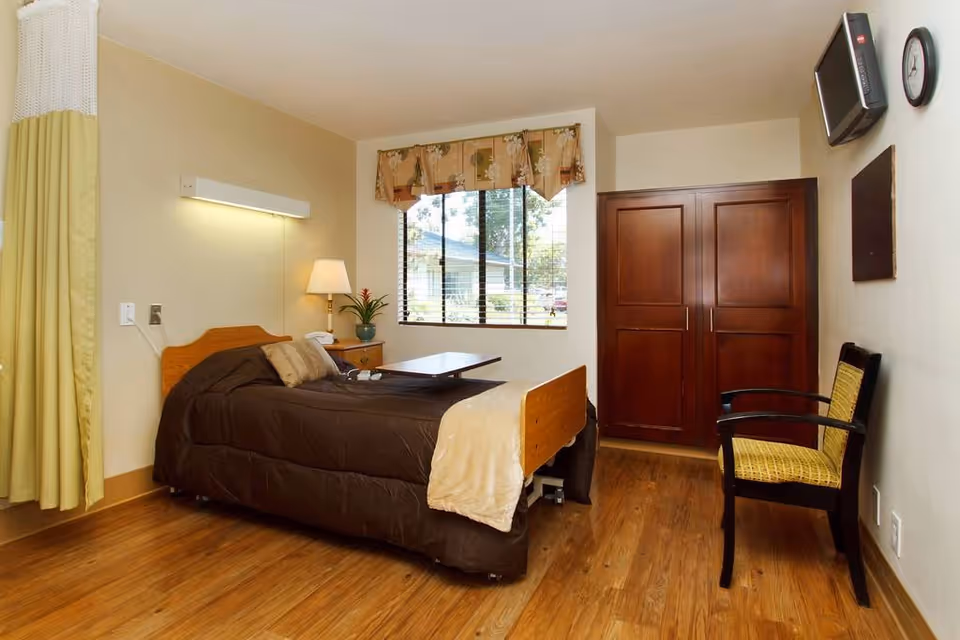 A neatly arranged bedroom in a healthcare facility featuring a single bed with brown bedding and a beige throw blanket. Next to the bed is a wooden nightstand with a lamp and a potted plant. A window with floral curtains lets in natural light. Across from the bed is a wooden wardrobe, a wall-mounted TV, a clock, and a chair with a yellow cushion. The room has wooden flooring and light-colored walls.