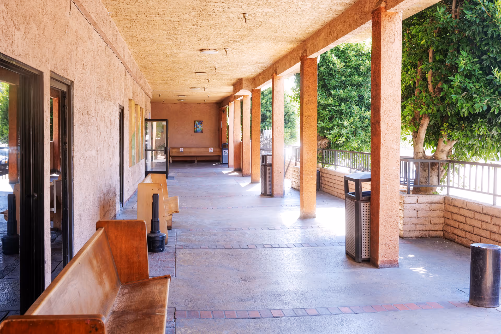 A covered outdoor walkway with wooden benches along the left side and trash bins on the right side. The walkway is supported by columns and overlooks a garden area with trees and a brick planter.
