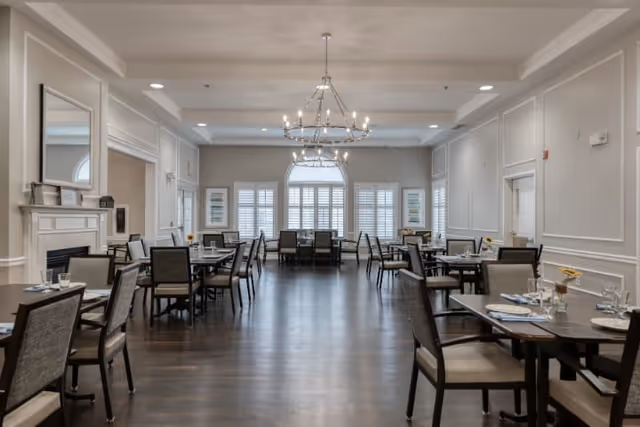 A spacious dining room with multiple tables and chairs arranged neatly. The room features large windows with white shutters, elegant chandeliers hanging from the ceiling, and light-colored walls with decorative molding. Tables are set with plates, glasses, and napkins, creating a welcoming atmosphere.
