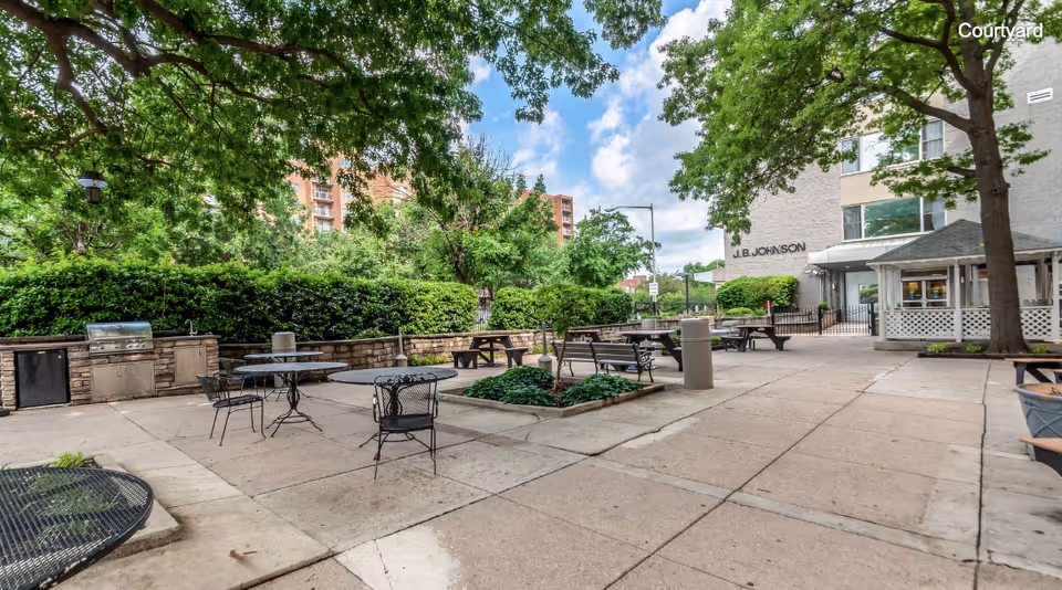 Outdoor courtyard area with several metal tables and chairs, picnic tables, a built-in grill station, and a small gazebo. The area is surrounded by trees and bushes, with a multi-story building labeled 'J.B. Johnson' in the background under a partly cloudy sky.