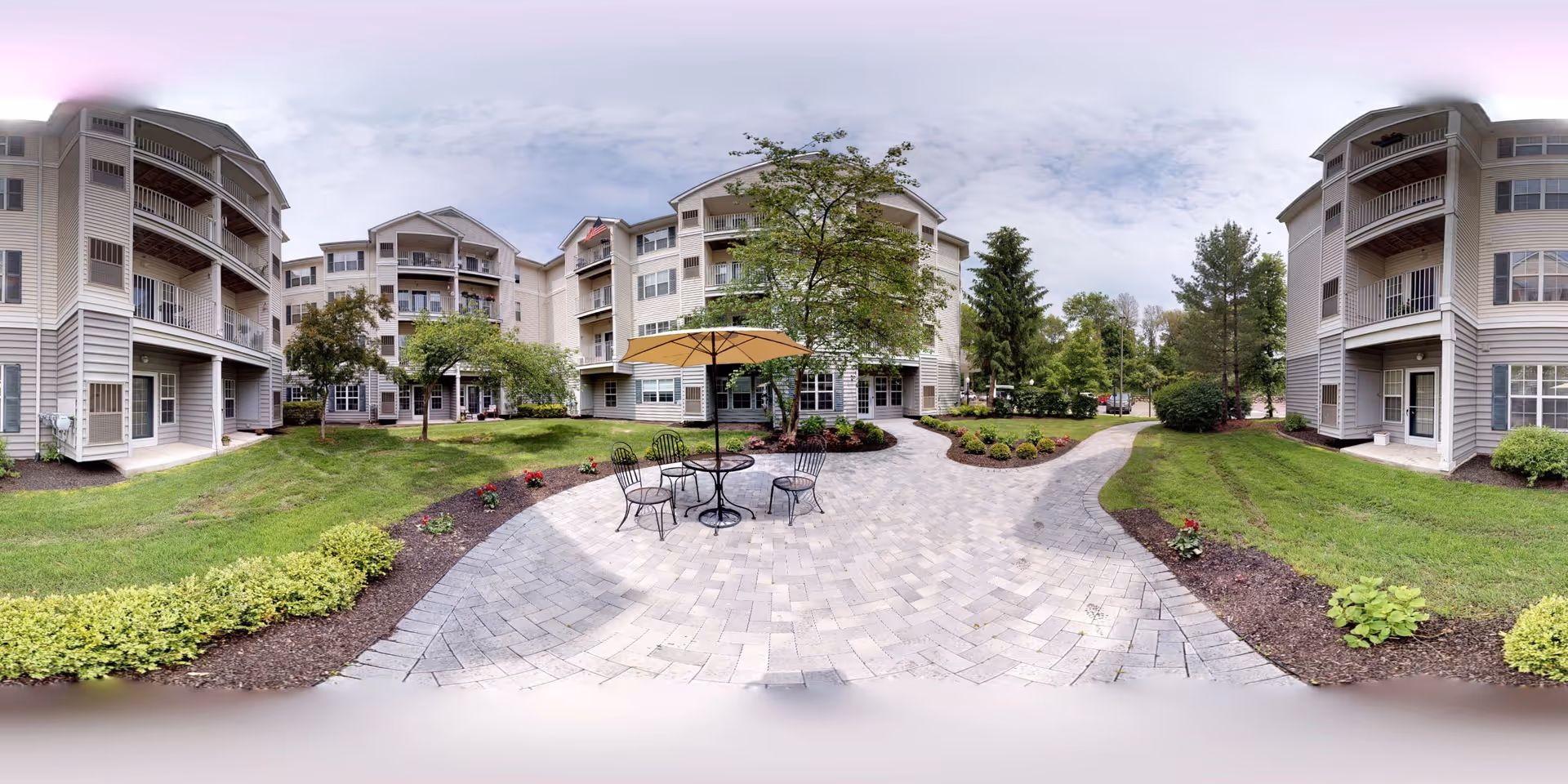 Central paved courtyard with a table, chairs and umbrella, surrounded by multi-story senior living buildings and landscaped lawns.