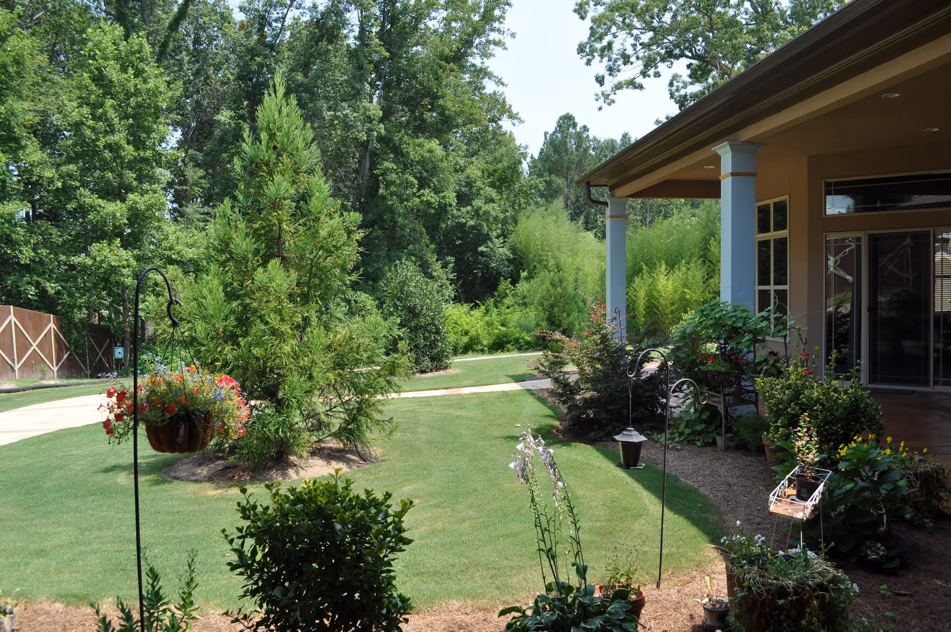A well-maintained garden area with green grass, various shrubs, and trees. There are hanging flower baskets and potted plants near a covered patio attached to a building. The scene is bright and sunny with a pathway leading through the garden.