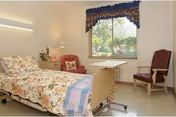A well-lit bedroom in a senior living facility featuring a hospital-style bed with floral bedding and a blue blanket, a pink armchair with a pillow, a wooden side table with a lamp and flowers, a window with blinds and a decorative valance, and a wooden chair with armrests.