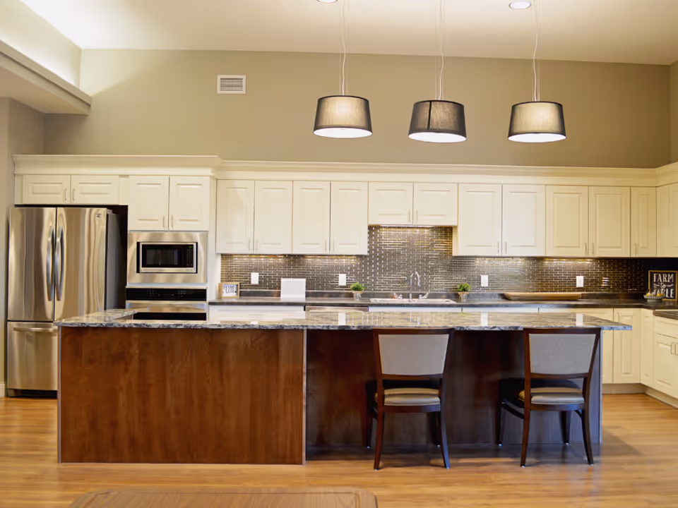 A modern kitchen with white cabinets, a stainless steel refrigerator, built-in microwave, and oven. There is a large island with a granite countertop and two chairs. Three pendant lights hang above the island, and the backsplash is made of small, dark tiles.