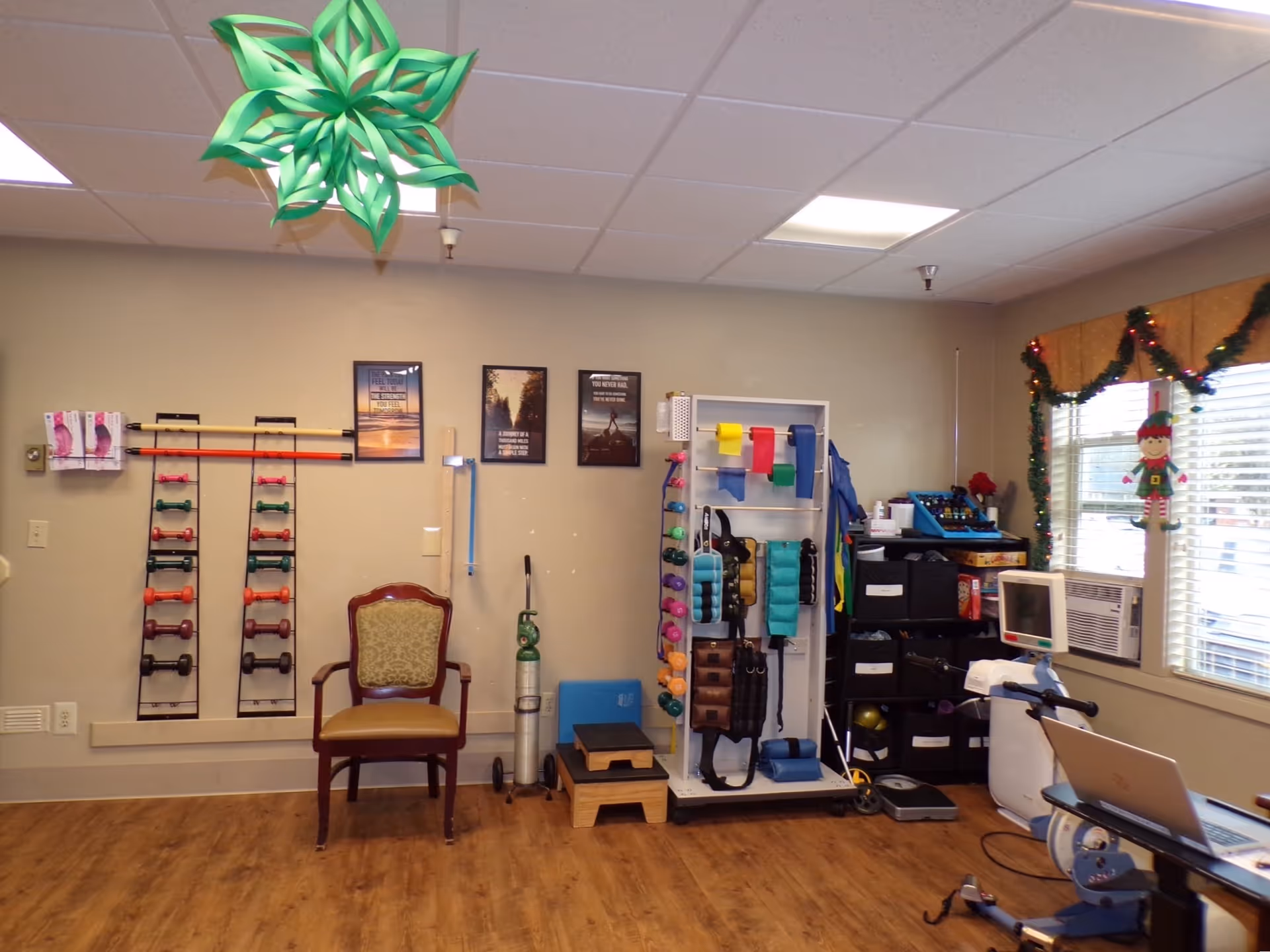 A rehabilitation or exercise room with various fitness equipment including dumbbells on wall racks, resistance bands, weights, and a stationary exercise bike. There is a wooden chair, a small step platform, and a window decorated with garland and a hanging elf ornament. The room has a wood floor and a drop ceiling with fluorescent lighting.