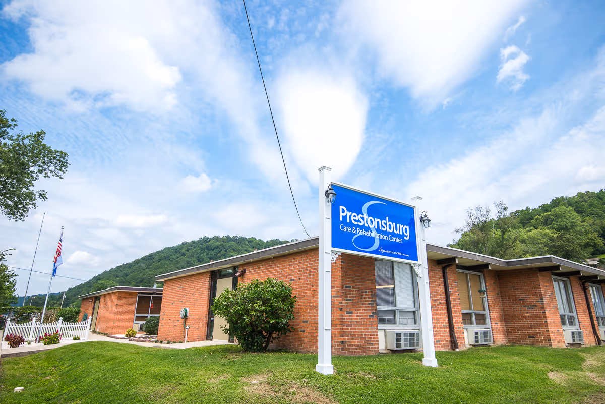 Exterior view of Prestonsburg Care & Rehabilitation Center, a single-story brick building with a blue sign in front. The building is surrounded by green grass and trees, with a partly cloudy sky overhead.