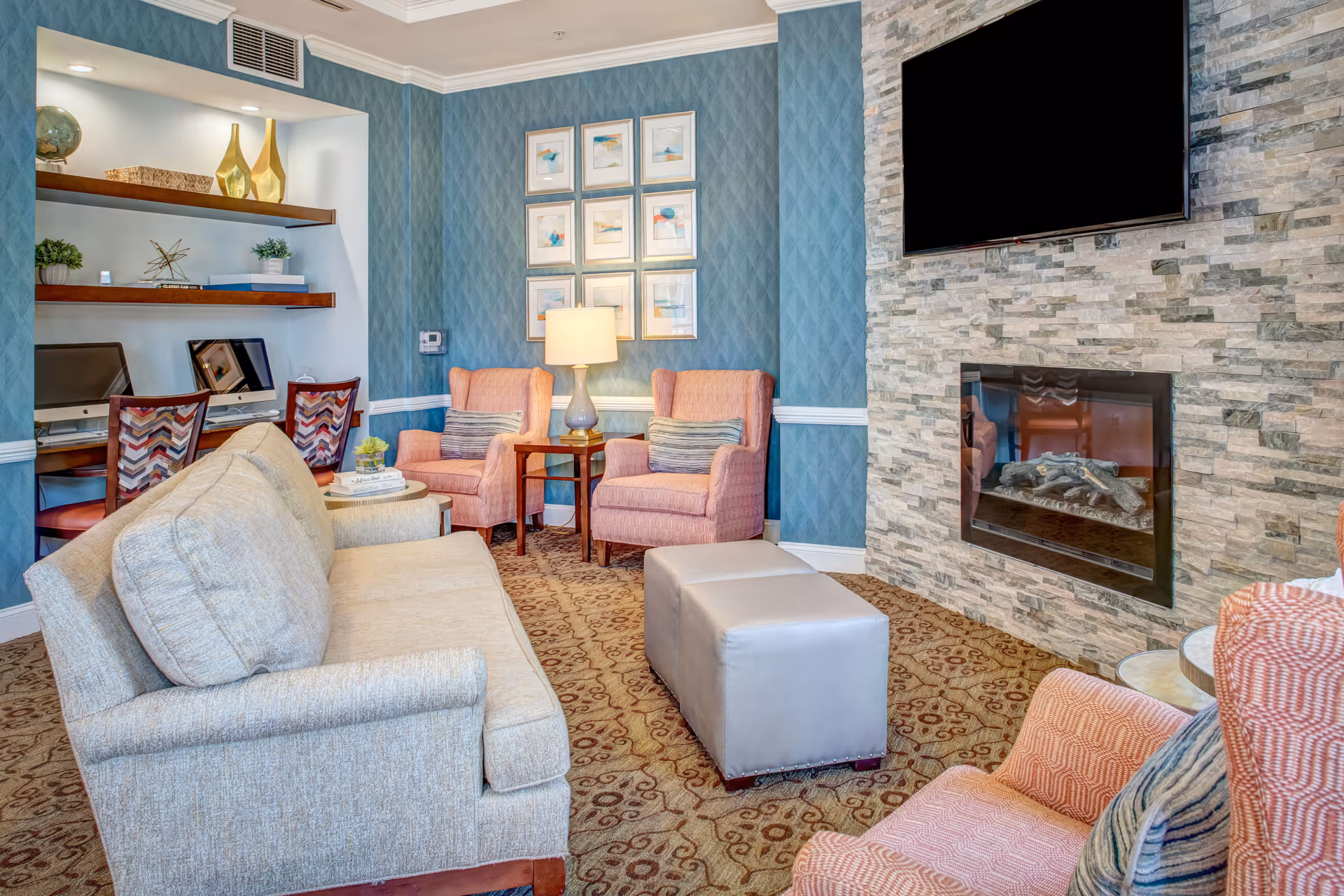 A cozy living room with a beige sofa, two pink armchairs with striped pillows, a small side table with a lamp, and a stone fireplace with a mounted flat-screen TV above it. There are two computer workstations with chairs and decorative shelves on the left side, and framed artwork on the blue patterned wall.