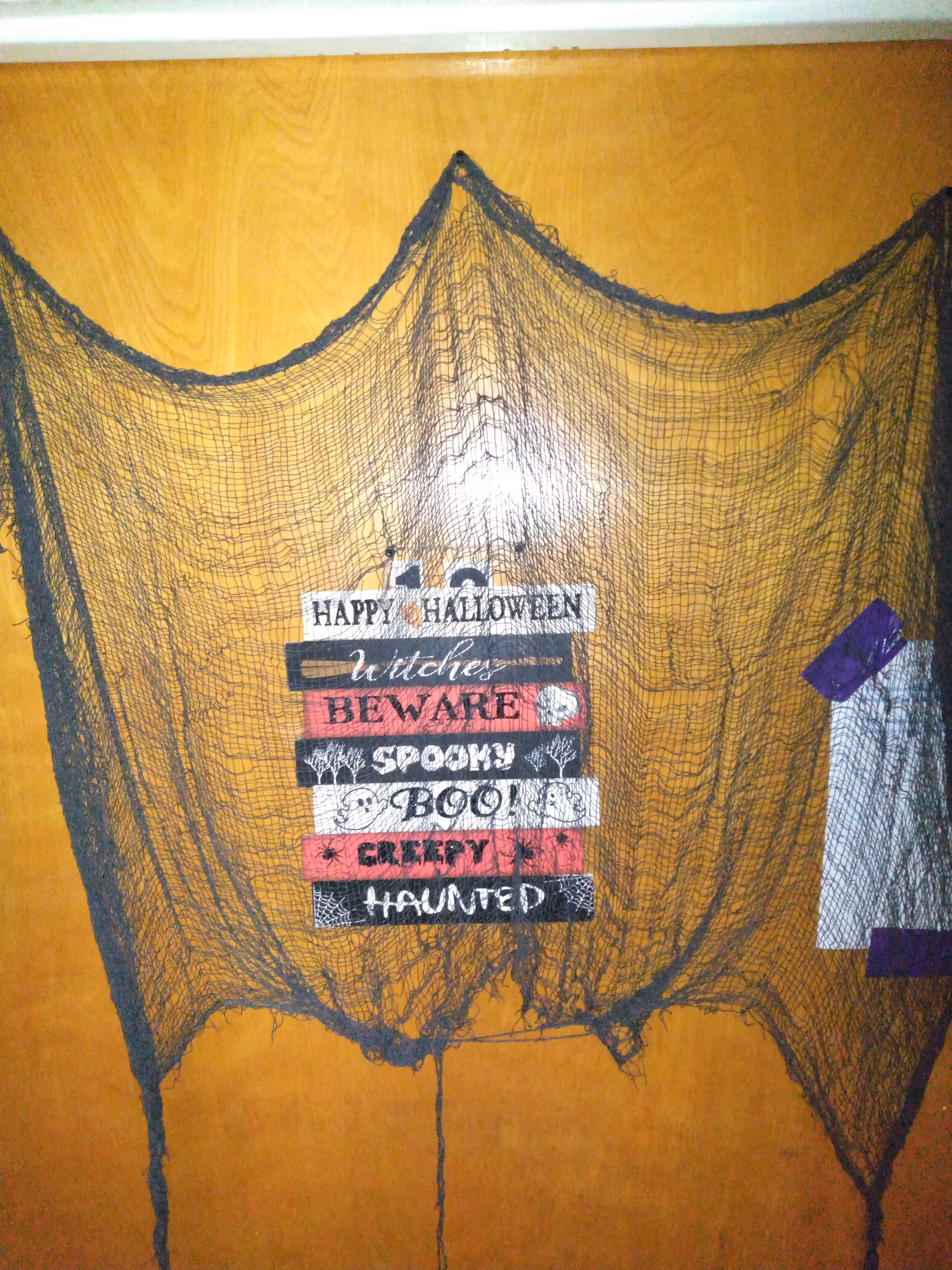 A wooden door decorated with a black spider web net and a Halloween-themed sign that reads 'Happy Halloween, Witches, Beware, Spooky, Boo, Creepy, Haunted'.