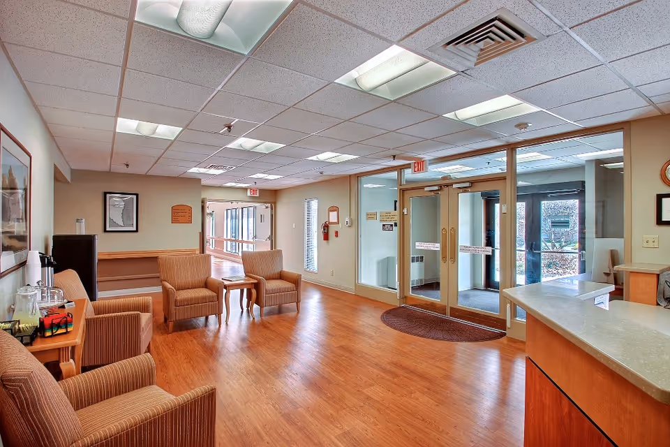 A waiting area inside Greenfield Healthcare Center featuring four striped armchairs arranged around a small wooden table. The room has wood flooring, a reception desk on the right, framed pictures on the walls, and large glass double doors leading outside. The ceiling has recessed fluorescent lighting and there is a water dispenser on the left side.