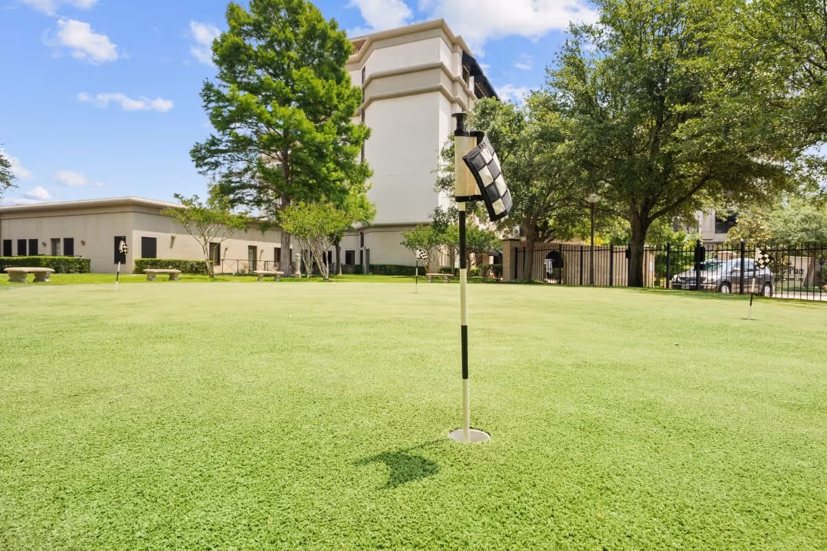 Outdoor putting green with several golf holes marked by black and white checkered flags, surrounded by trees, benches, and buildings under a blue sky with scattered clouds.