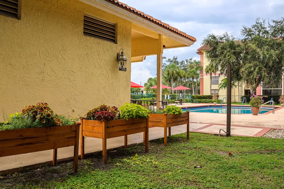 Outdoor area of a senior living facility featuring a beige stucco building wall with a lantern light and a restroom sign. In front of the building are three wooden planter boxes with various green and red plants. In the background, there is a swimming pool surrounded by a concrete deck, red umbrellas, palm trees, and a multi-story building.